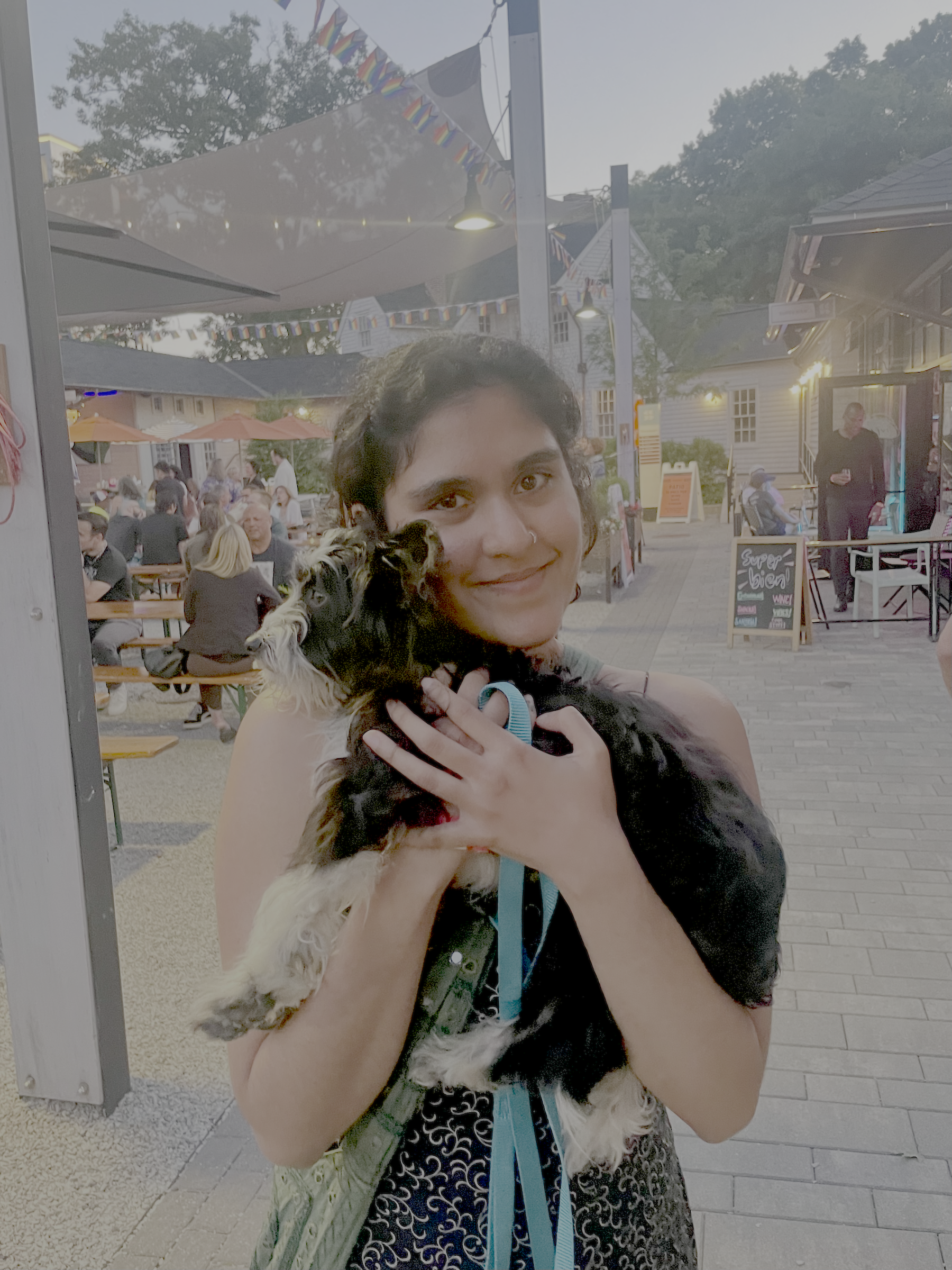 Vinitha, an Indian woman with tan skin, smiles at the camera holding a small black and white dog closer to her. She has black curly hair that is tied back and stands in front of a restaurant patio.